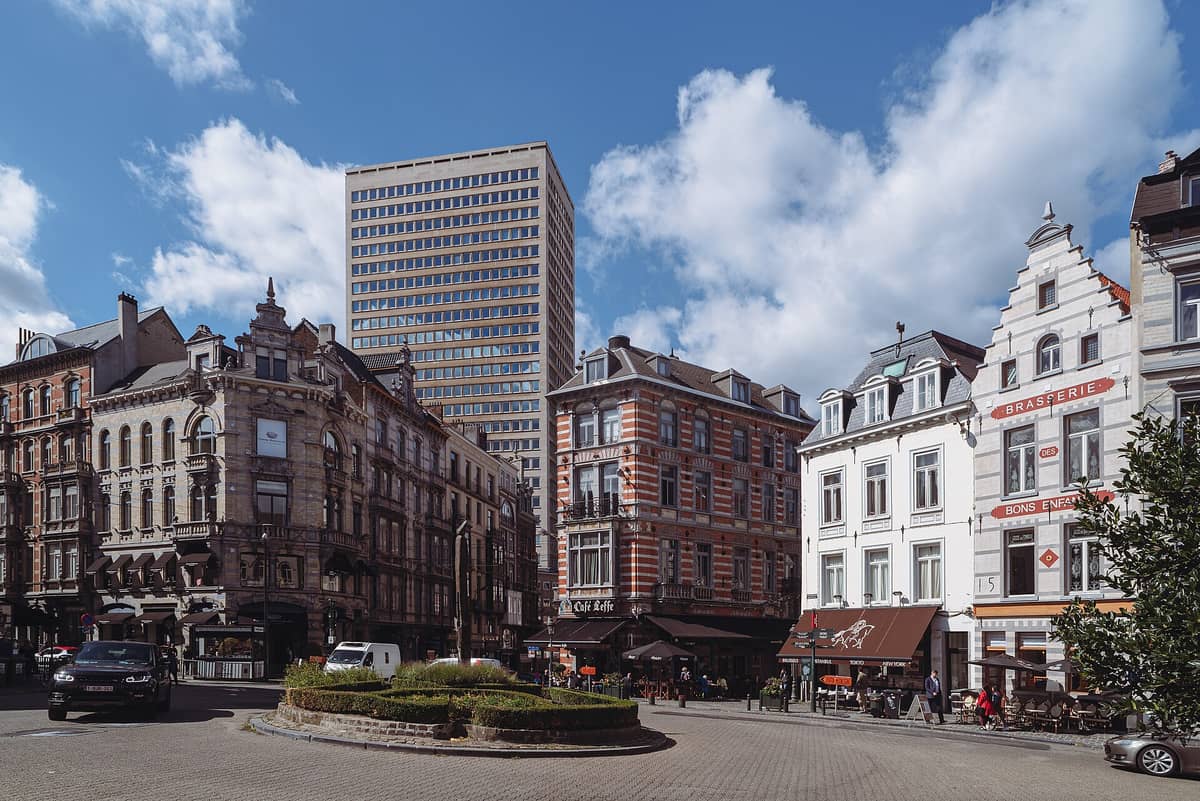 Vista da Place du Grand Sablon em Bruxelas com elegantes fachadas históricas, terraços de café animados e uma torre moderna ao fundo sob um céu azul.
