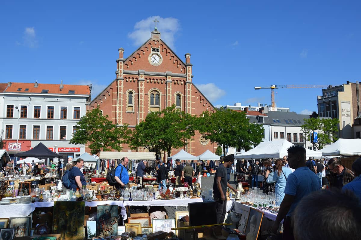 Dia movimentado na feira da ladra da Place du Jeu de Balle em Bruxelas, com bancas de antiguidades e uma igreja de tijolos vermelhos ao fundo.