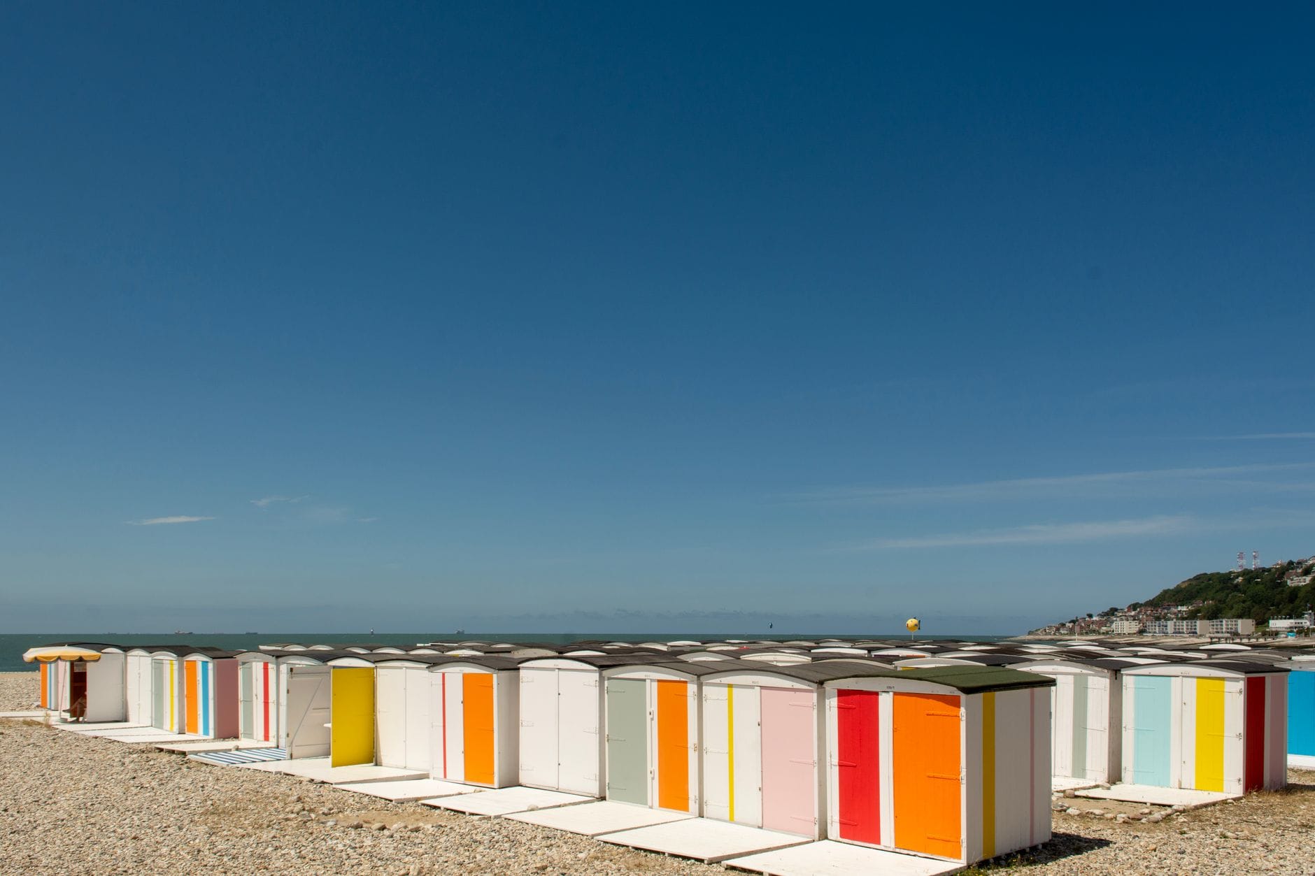 Plage du Havre'ın çakıllı kıyısında sıralanan renkli çizgili plaj kabinleri; mavi gökyüzü ve uzakta denizle buluşan kayalıklar.