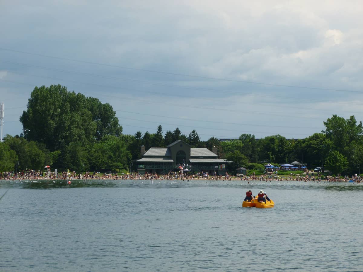 Blick auf die Plage Jean-Doré auf der Île Notre-Dame in Montreal mit belebtem Sandstrand, See und Menschen auf einem gelben Tretboot im Vordergrund.