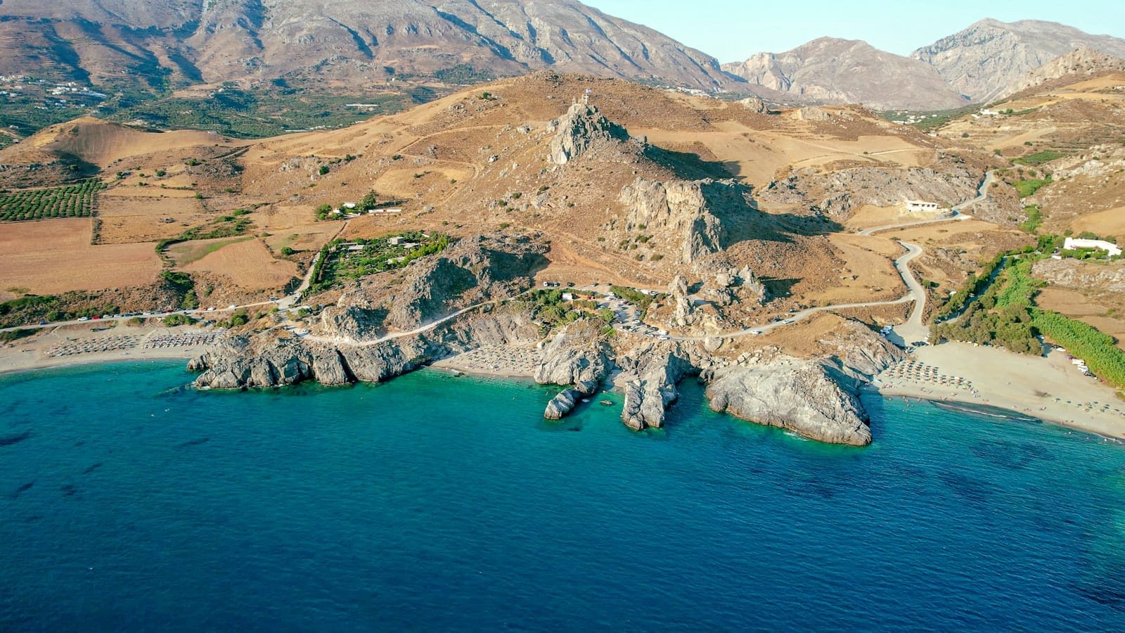 Vista aerea della spiaggia di Plakias sulla costa meridionale di Creta, con acqua turchese, una mezzaluna di sabbia, lettini e montagne aspre sullo sfondo.