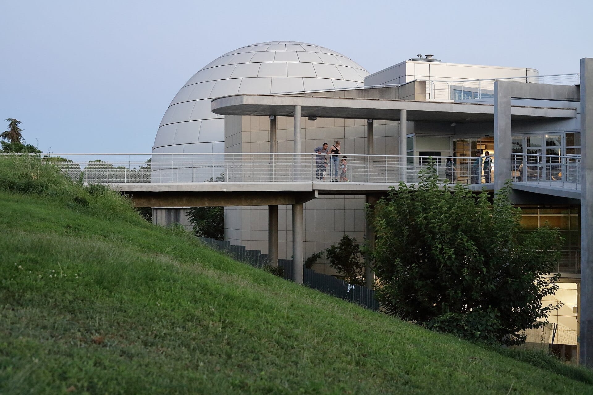 Esterno moderno del Planetario de Madrid con la sua caratteristica cupola, una collina verde erbosa in primo piano e visitatori che godono della serata.