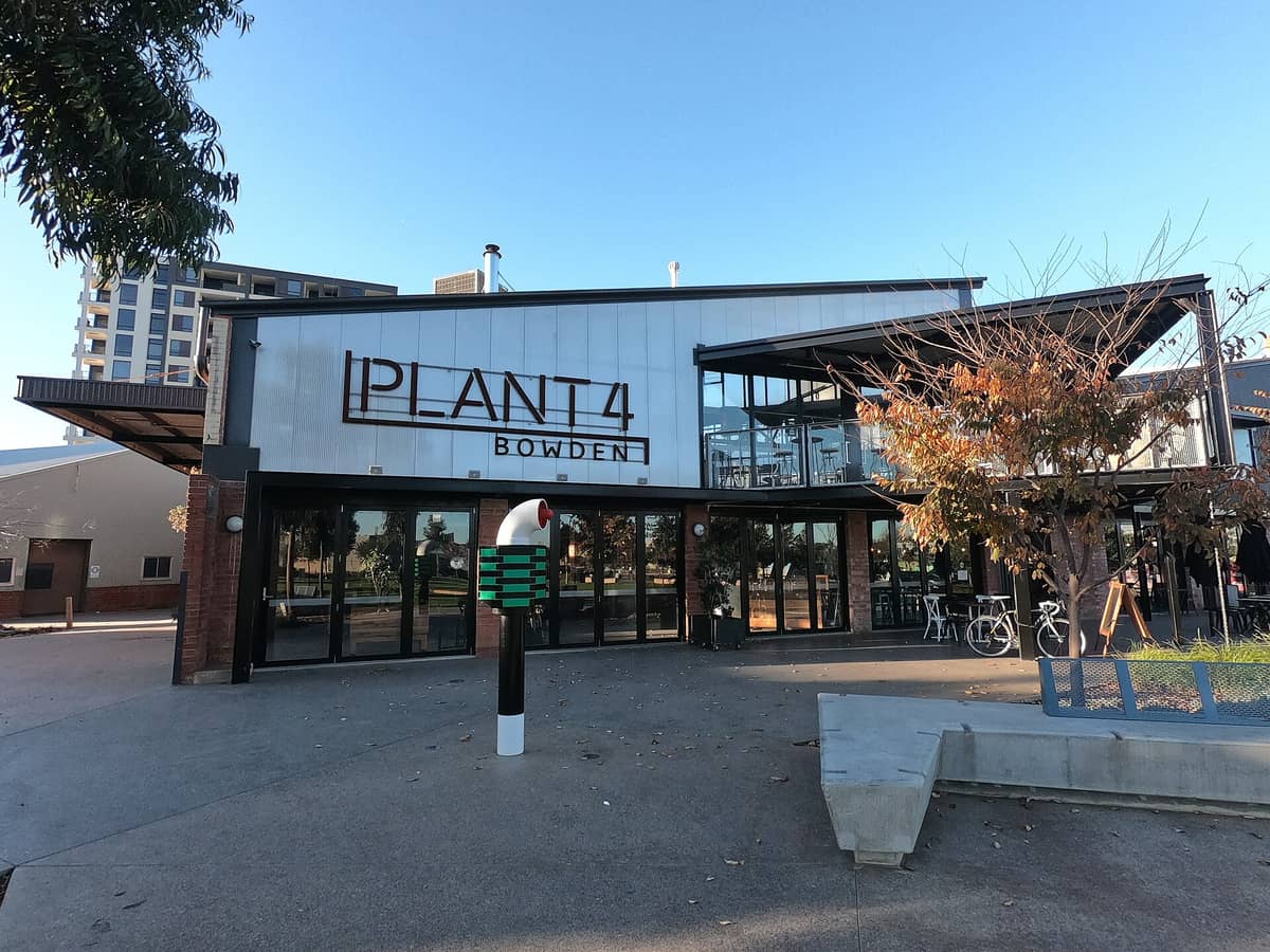 Exterior view of Plant 4 Bowden, with large sign on modern industrial building, outdoor seating, and trees in front on a clear day.