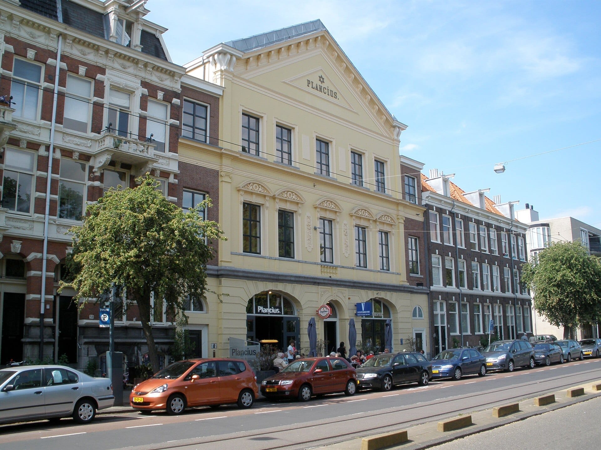 Histórico edificio Plancius con ornamentada fachada amarilla y adyacentes casas señoriales holandesas clásicas en el barrio de Plantage y el Barrio Judío, Ámsterdam, bajo un cielo azul despejado.