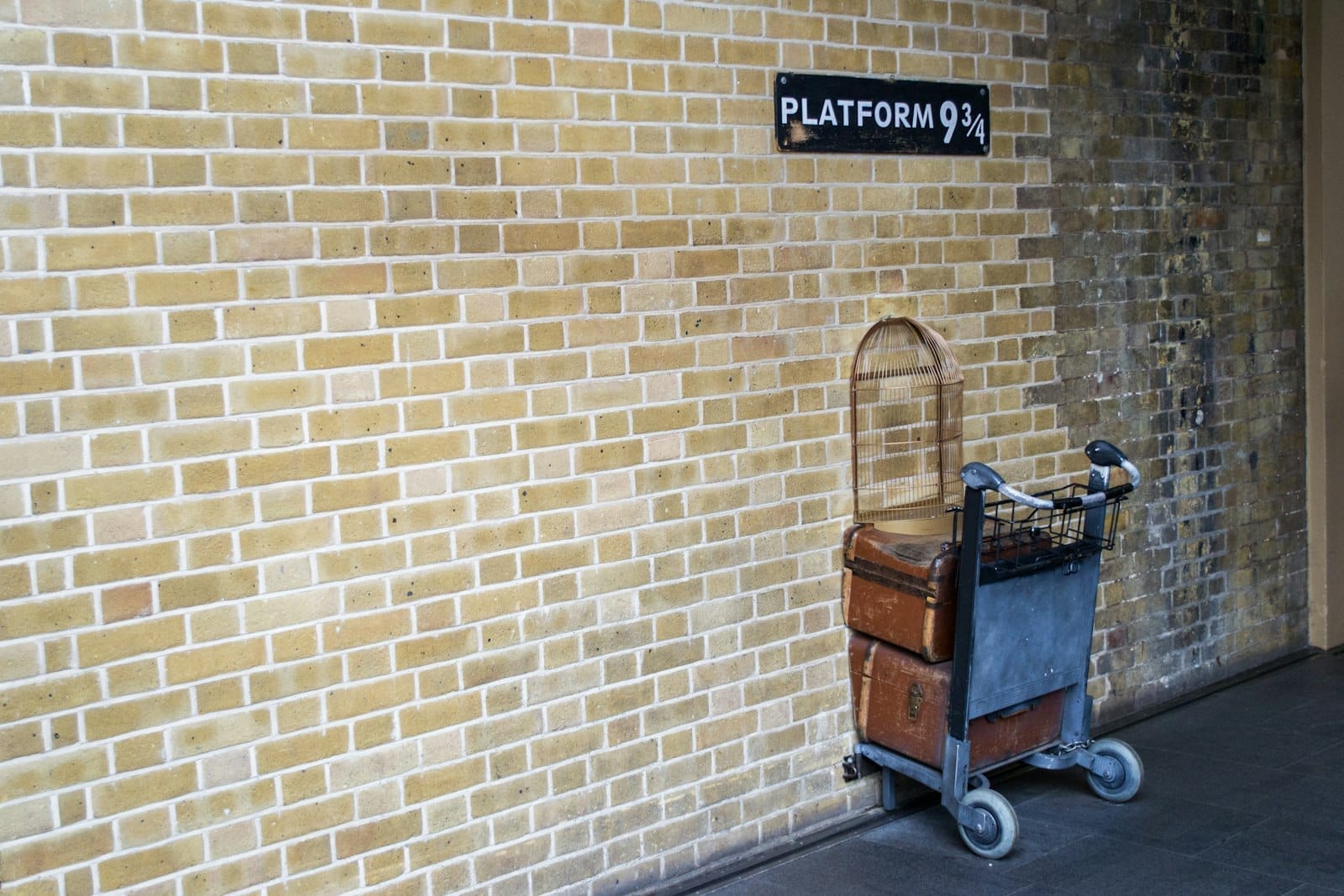 Un carrello portabagagli con valigie vintage e una gabbia per uccelli appare a metà nel muro di mattoni sotto il cartello del Binario 9¾ a King's Cross.