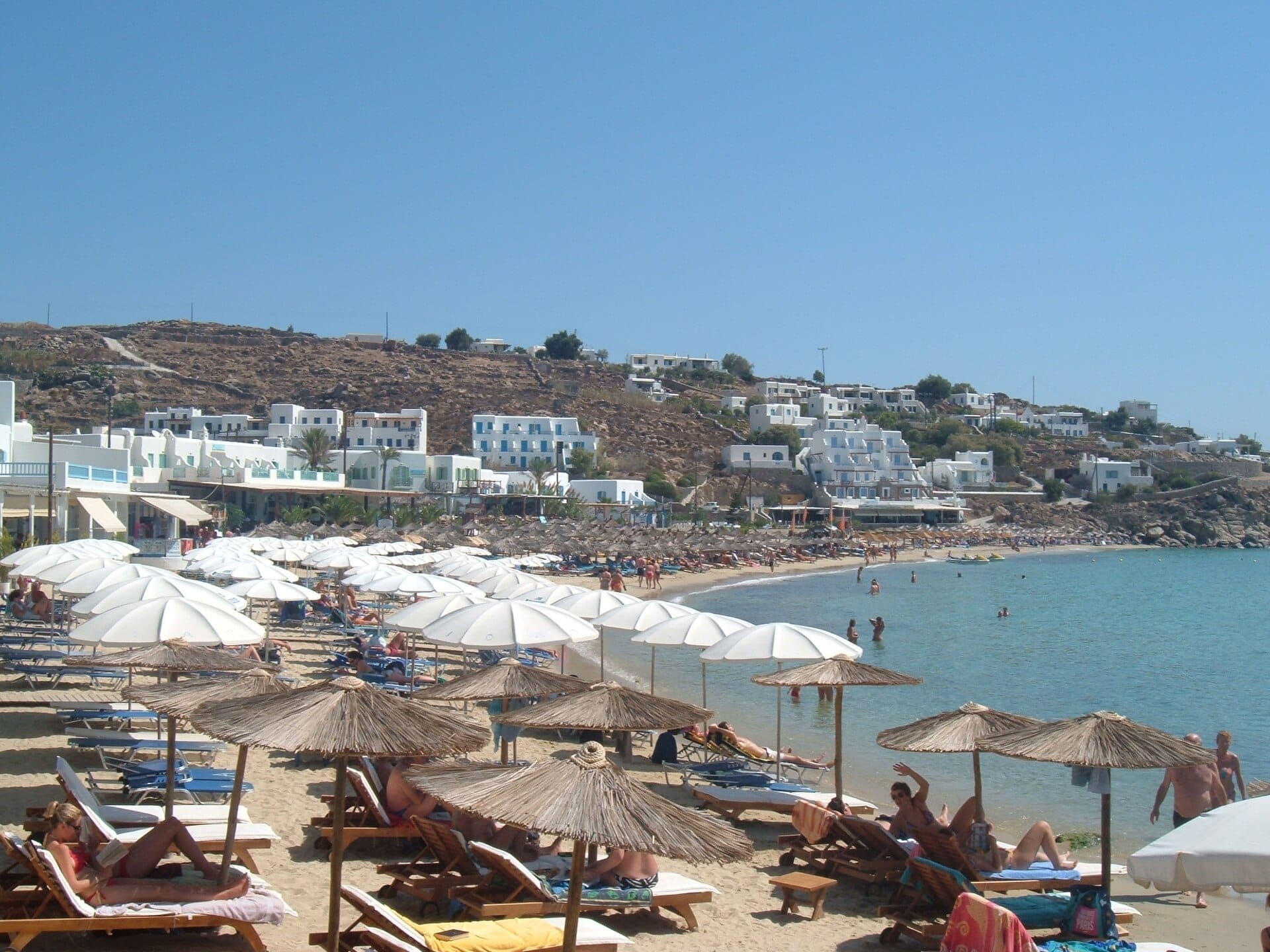Playa Platis Gialos con filas de tumbonas y sombrillas sobre arena dorada, agua azul cristalina y hoteles encalados en la ladera de Mykonos.