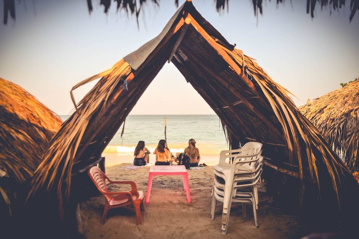 View from inside a rustic palm hut at Playa Blanca, Isla Barú, showing three people sitting on the sand facing turquoise water.