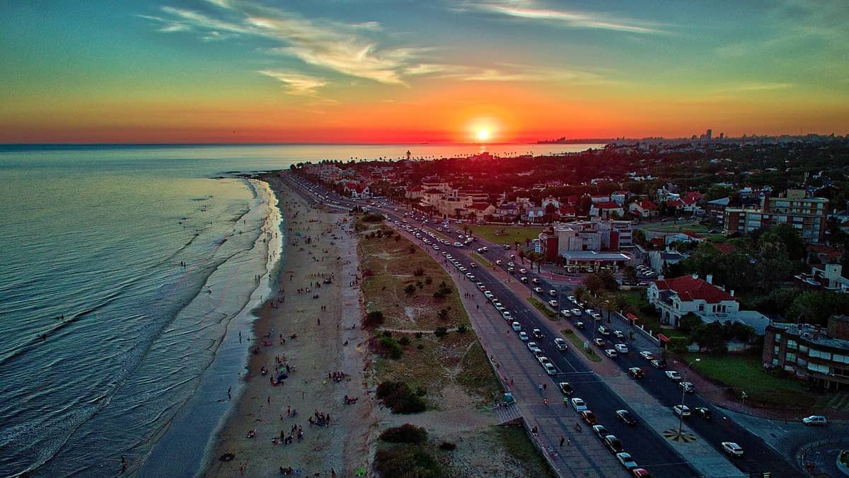 มุมมองทางอากาศของ Playa Carrasco ยามพระอาทิตย์ตกดิน แสดงให้เห็นคลื่นสงบ ชายหาดทราย นักท่องเที่ยวประปราย บ้านเรือนที่พักอาศัย และถนนชายฝั่งที่คึกคัก
