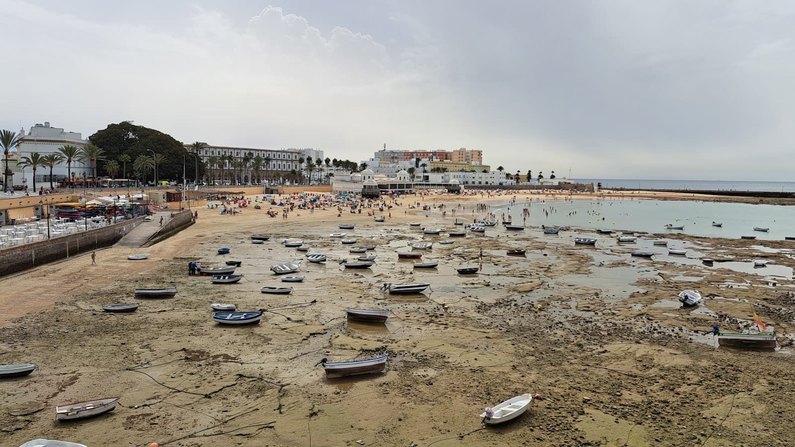 Wide view of Playa de La Caleta at low tide, with small boats resting on wet sand and the historic walled city, palm trees, and beachgoers in the background.