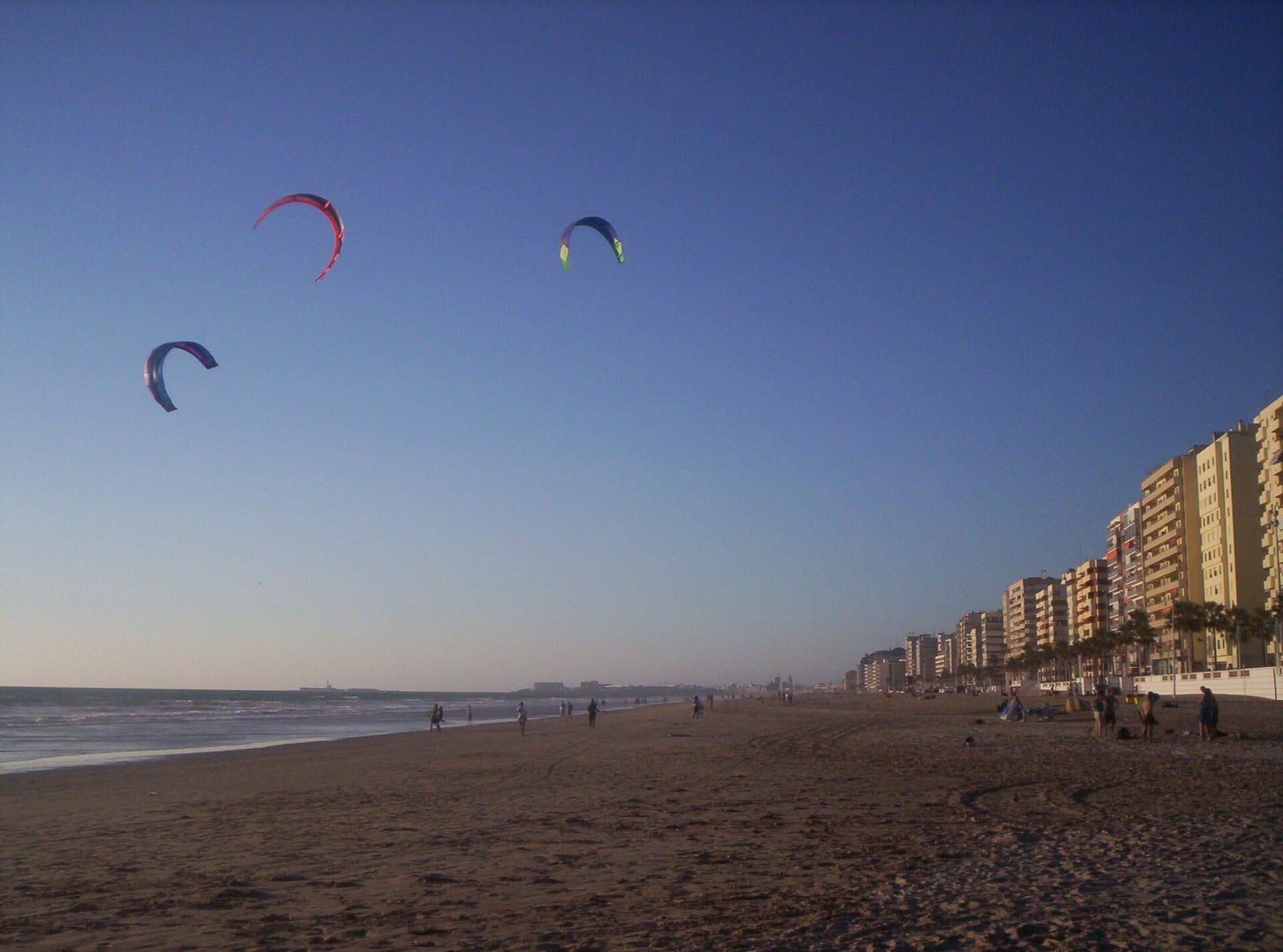 Wide sandy Playa de la Victoria with several people walking, tall apartment buildings along the promenade, and colorful kitesurfers in a clear blue sky.