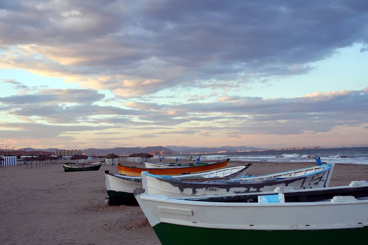 Un'ampia spiaggia sabbiosa alla Playa de las Arenas di Valencia con colorate barche da pesca sulla riva, edifici della città sullo sfondo e un cielo al tramonto.