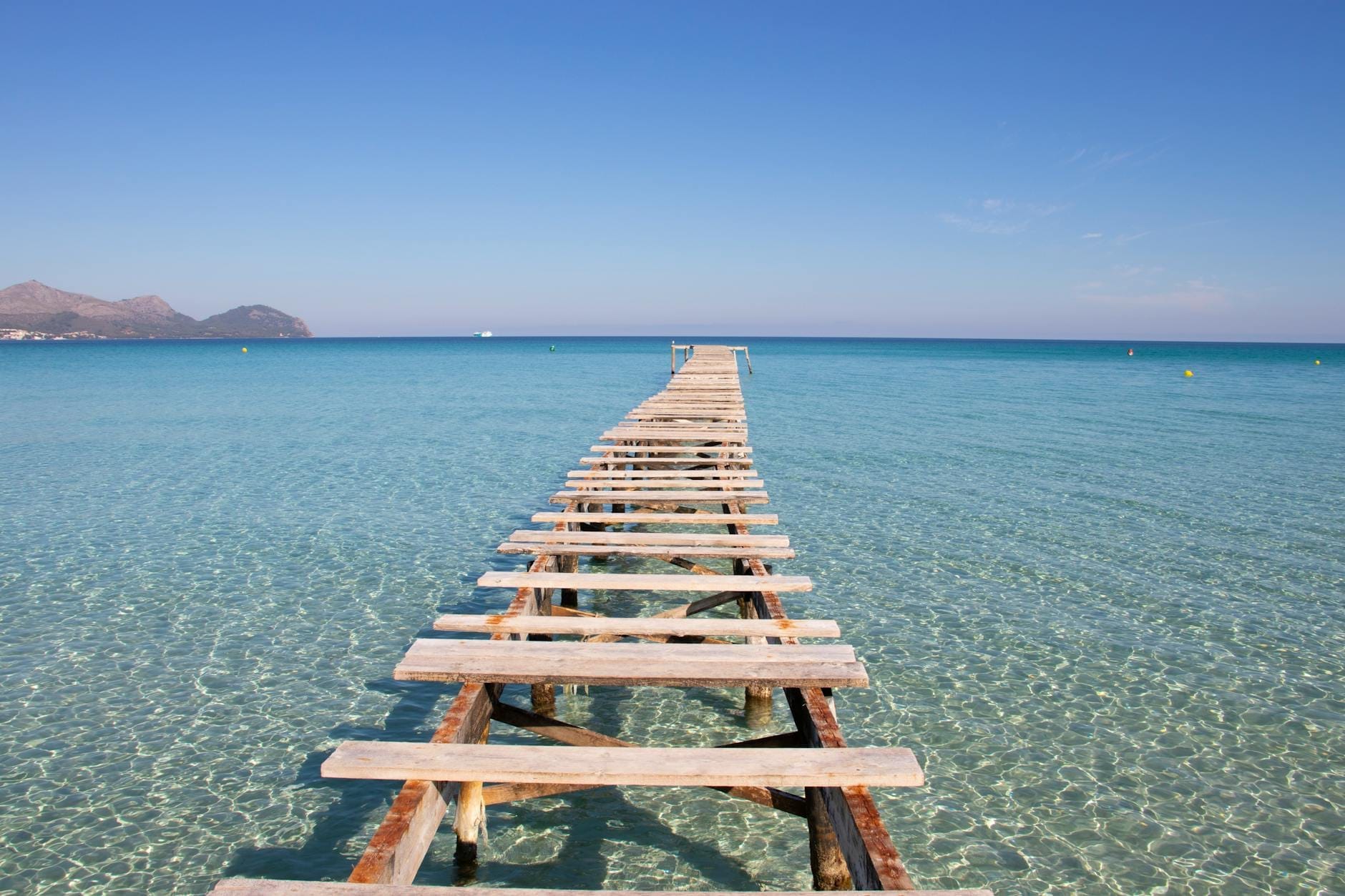Acqua turchese cristallina e un rustico pontile in legno che si allunga verso il mare a Playa de Muro, con le montagne sullo sfondo sotto un cielo azzurro.