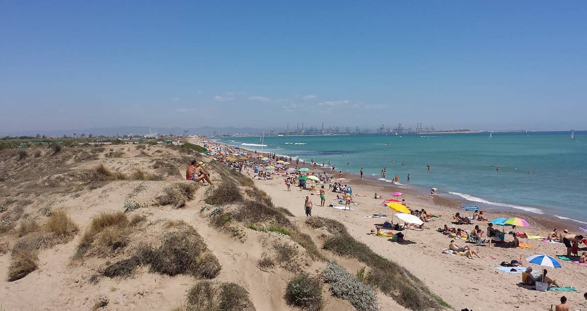 Vista della Playa de Pinedo, Valencia, con dune naturali sabbiose, acqua turchese e persone che si rilassano sotto gli ombrelloni; sullo sfondo si intravede il porto.