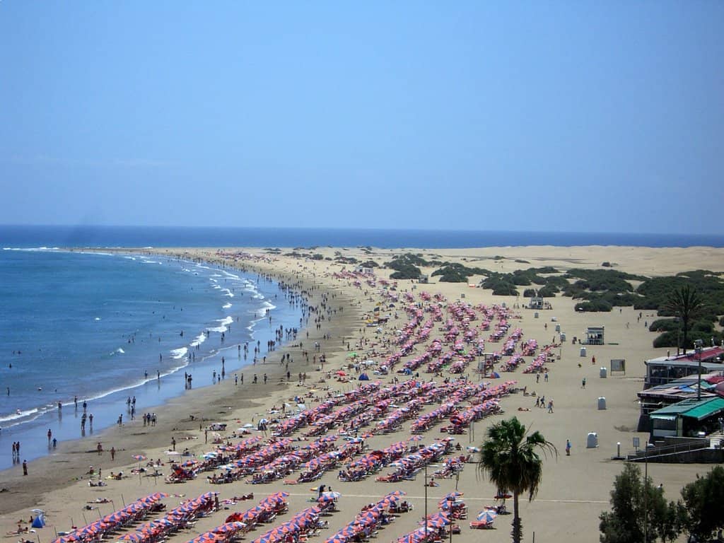 Широкий вид на пляж Playa del Inglés с рядами разноцветных шезлонгов, мягкими волнами и золотым песком, уходящим к дюнам Maspalomas.
