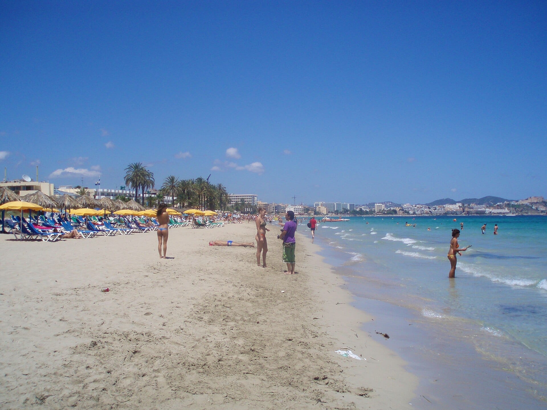 Vista ampia della spiaggia di Playa d'en Bossa con lettini, ombrelloni gialli, palme e persone che si godono la riva sotto un cielo azzurro limpido.