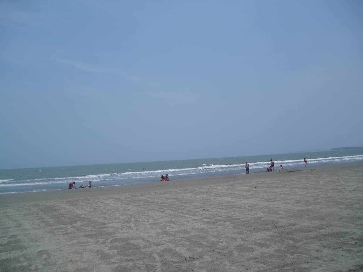 Wide sandy shore of Playa Marbella in Cartagena with scattered people near the water, gentle waves, and a clear blue sky above.