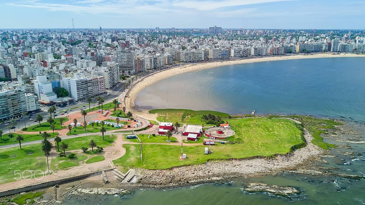 ภาพถ่ายทางอากาศของชายหาด Playa Pocitos ที่เห็นแนวทรายโค้ง ทางเดินริม Rambla สวนเขียวขจี และเส้นขอบฟ้าเมืองมอนเตวิเดโอใต้ท้องฟ้าแจ่มใส