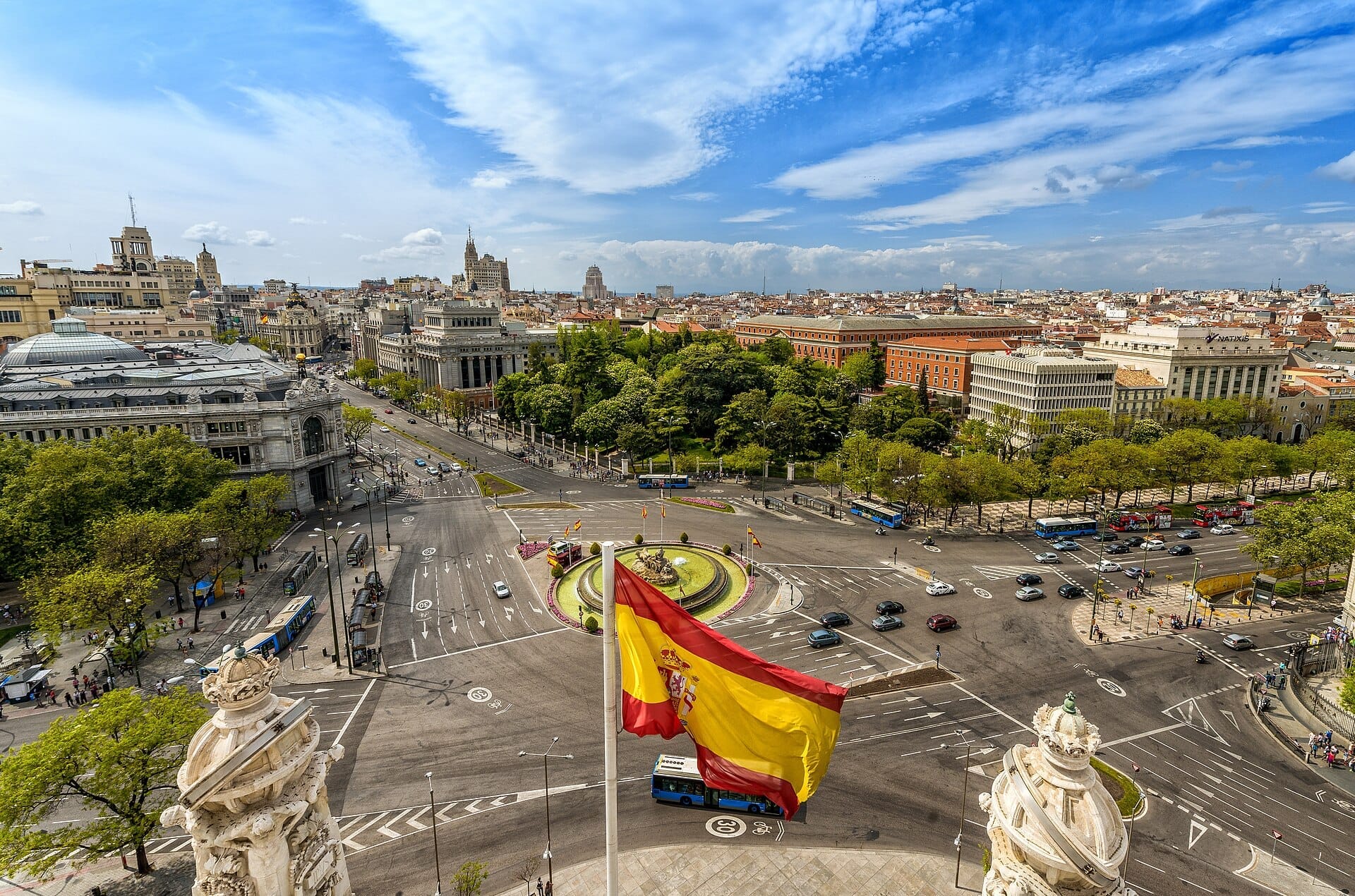 Vista aerea di Plaza de Cibeles a Madrid con la fontana iconica, edifici storici, rigogliosa vegetazione e una bandiera spagnola sventolante sotto cieli azzurri.