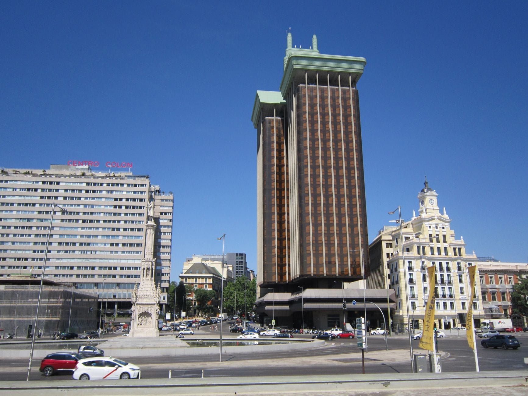 Vista panoramica di Plaza de Colón a Madrid con il monumento a Colombo, le torri moderne, gli edifici circostanti e la piazza aperta sotto un cielo azzurro limpido.