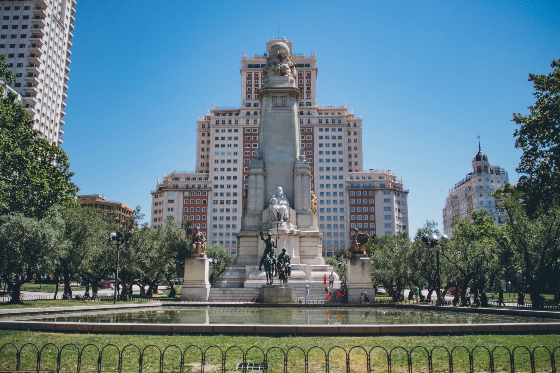 Vista ampia di Plaza de España a Madrid con il prominente monumento a Cervantes, il verde rigoglioso e gli edifici storici circostanti sotto un cielo azzurro terso.