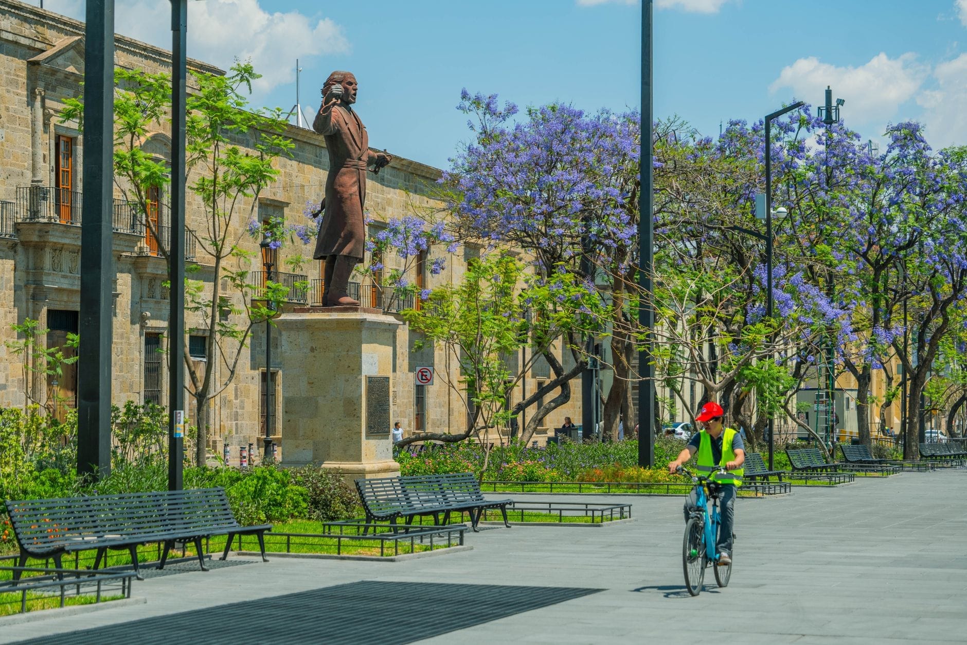 Vista della Plaza de la Liberación con una statua, panchine, jacarande in fiore e un ciclista che attraversa la piazza pedonale nel centro storico di Guadalajara.