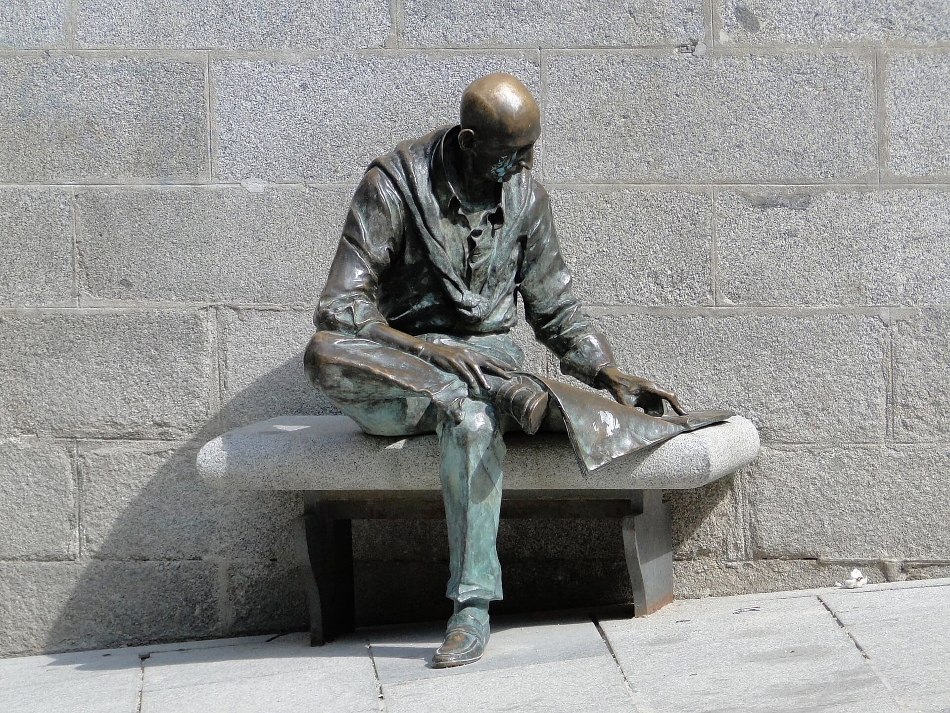 Statua in bronzo di un uomo seduto che legge su una panchina di pietra in Plaza de la Paja, con un muro di pietra grigia dalla superficie irregolare sullo sfondo.