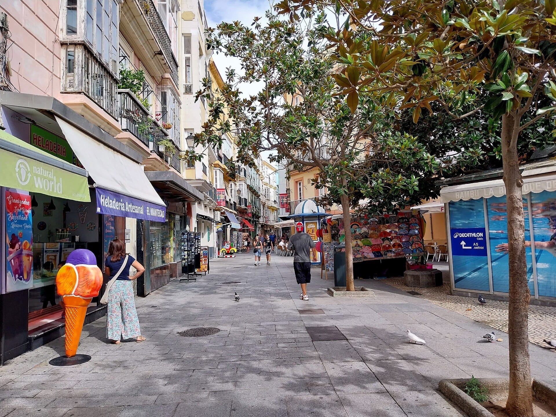 Strada pedonale in Plaza de las Flores, Cádiz, con colorata gelateria, bancarella di ombrelli, alberi ed edifici storici alla luce del giorno.