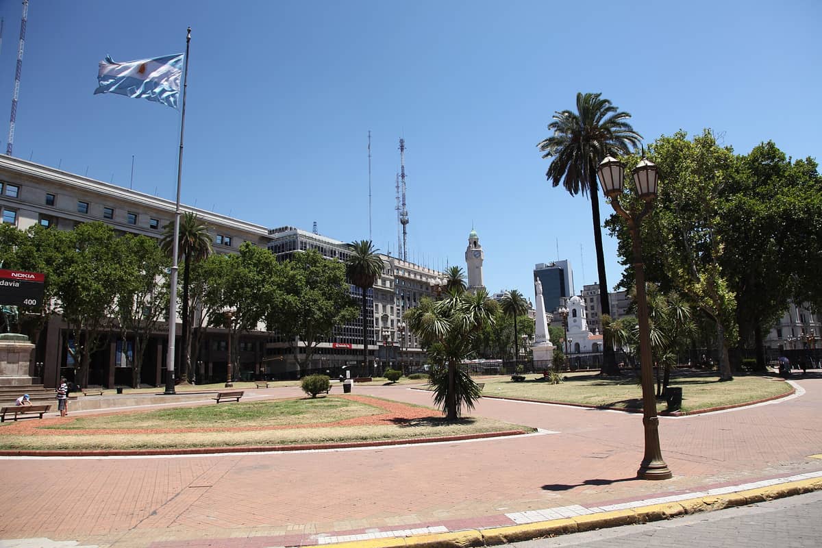 มุมมองกว้างของ Plaza de Mayo ในบัวโนสไอเรส เห็นจัตุรัสโล่ง ธงชาติ ที่นั่ง ต้นปาล์ม และอาคารประวัติศาสตร์เป็นฉากหลังท่ามกลางวันที่แดดสดใส