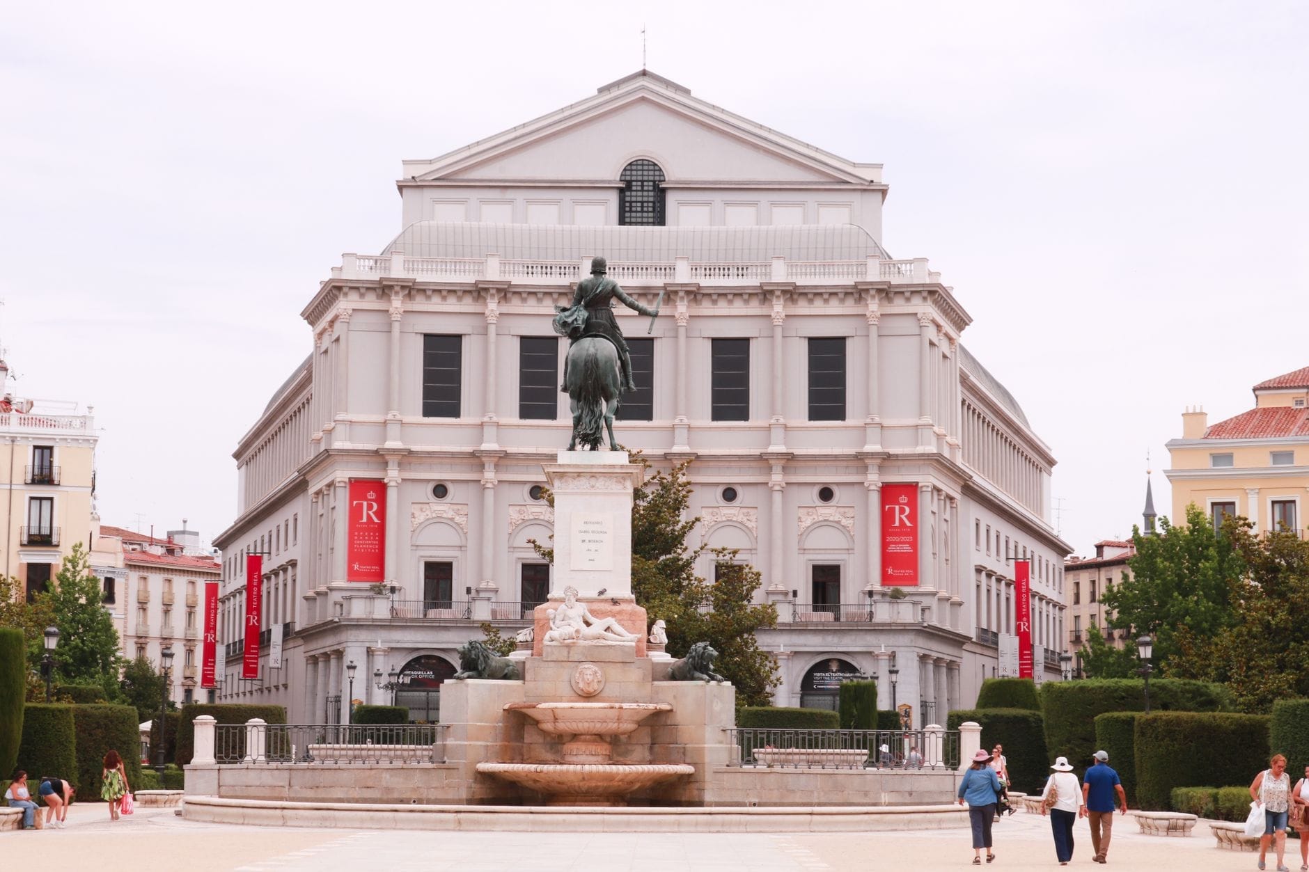 Vista panoramica di Plaza de Oriente a Madrid con statua e fontana al centro, il Teatro Real sullo sfondo e persone che passeggiano nella elegante piazza.