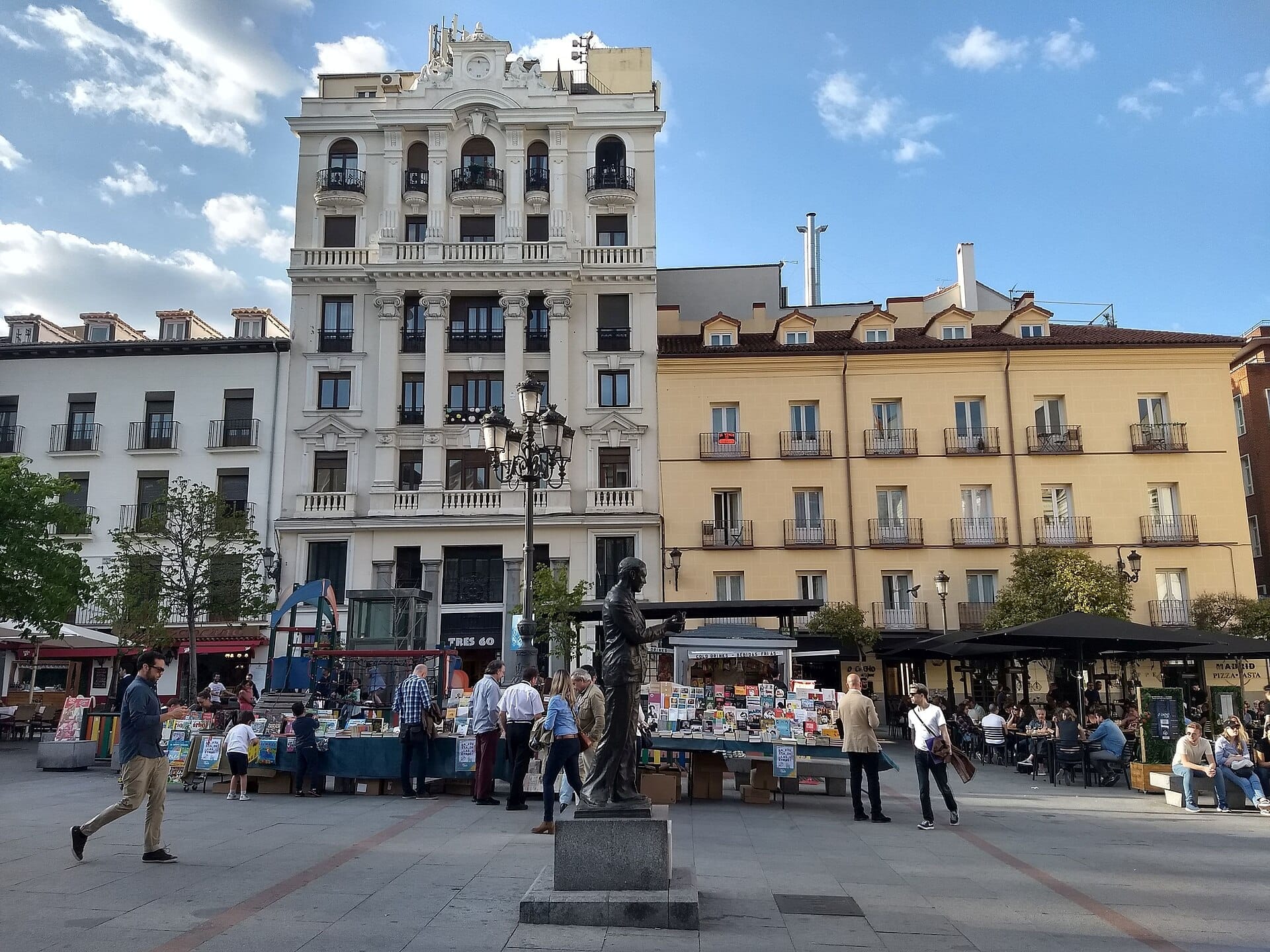 Plaza de Santa Ana a Madrid con edifici storici, una statua centrale, bancarelle di libri all'aperto e persone che godono delle terrazze dei caffè in una giornata di sole.