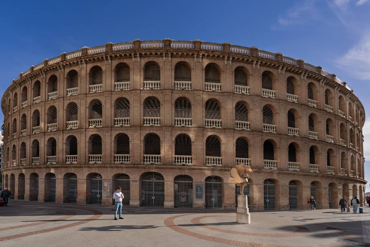Vista grandangolare della facciata neoclassica della Plaza de Toros de Valencia sotto un cielo blu, con i dettagli ad arco dell'arena e la piazza antistante.