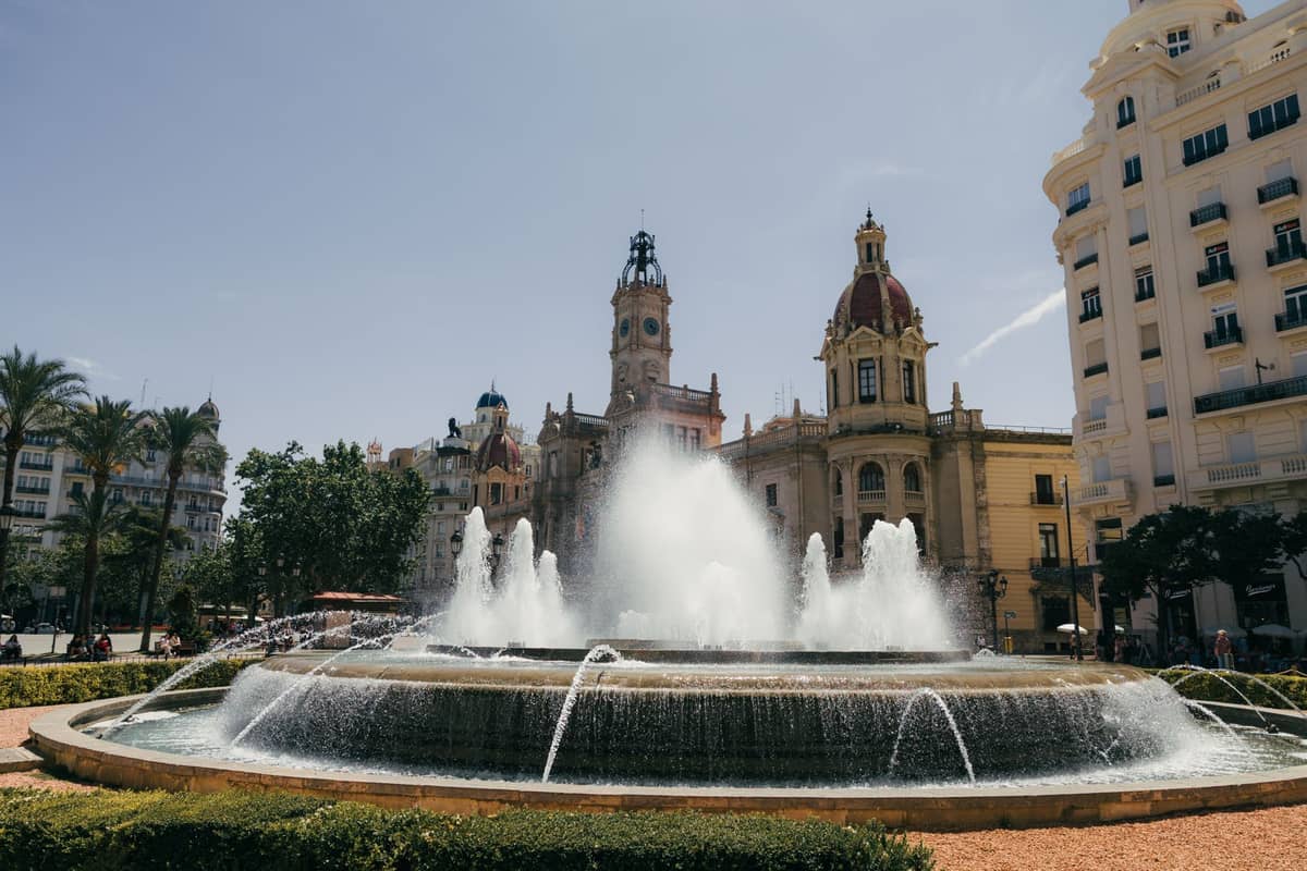 Vista ampia della Plaza del Ayuntamiento di Valencia con la fontana centrale in funzione, circondata dagli edifici storici tra cui il Municipio sotto un cielo azzurro limpido.