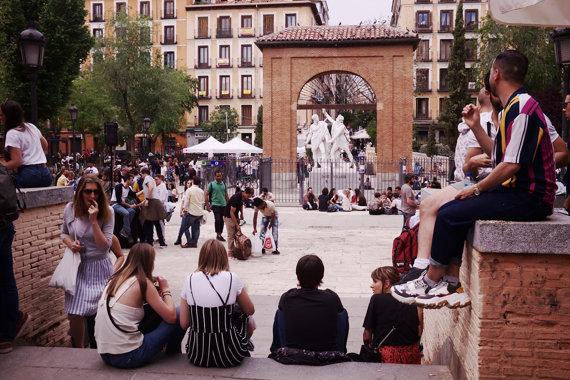Vista della Plaza del Dos de Mayo a Madrid, con persone che si rilassano intorno all'arco centrale e alle statue, circondata da colorati edifici residenziali.