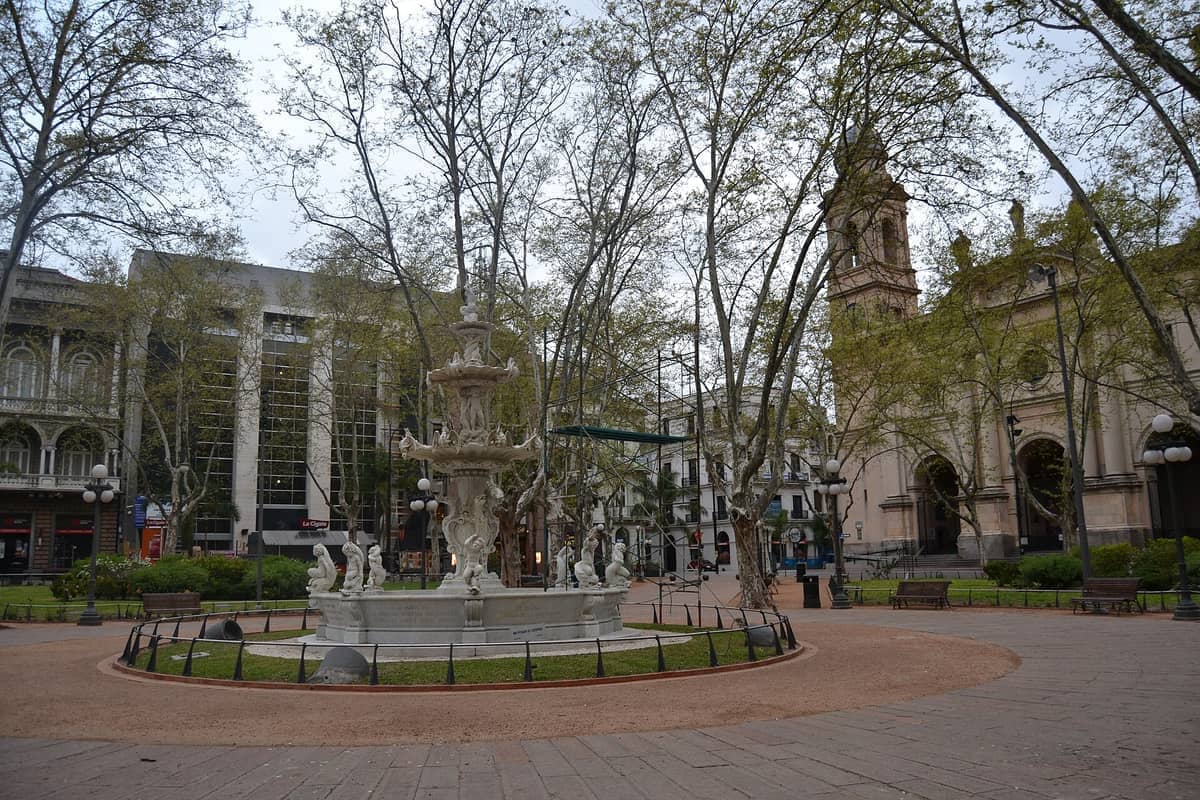 น้ำพุกลางจัตุรัสล้อมรอบด้วยม้านั่ง ต้นไม้ใหญ่ และอาคารประวัติศาสตร์ที่ Plaza Matriz เมืองมอนเตวิเดโอ ใต้ท้องฟ้ามีเมฆ