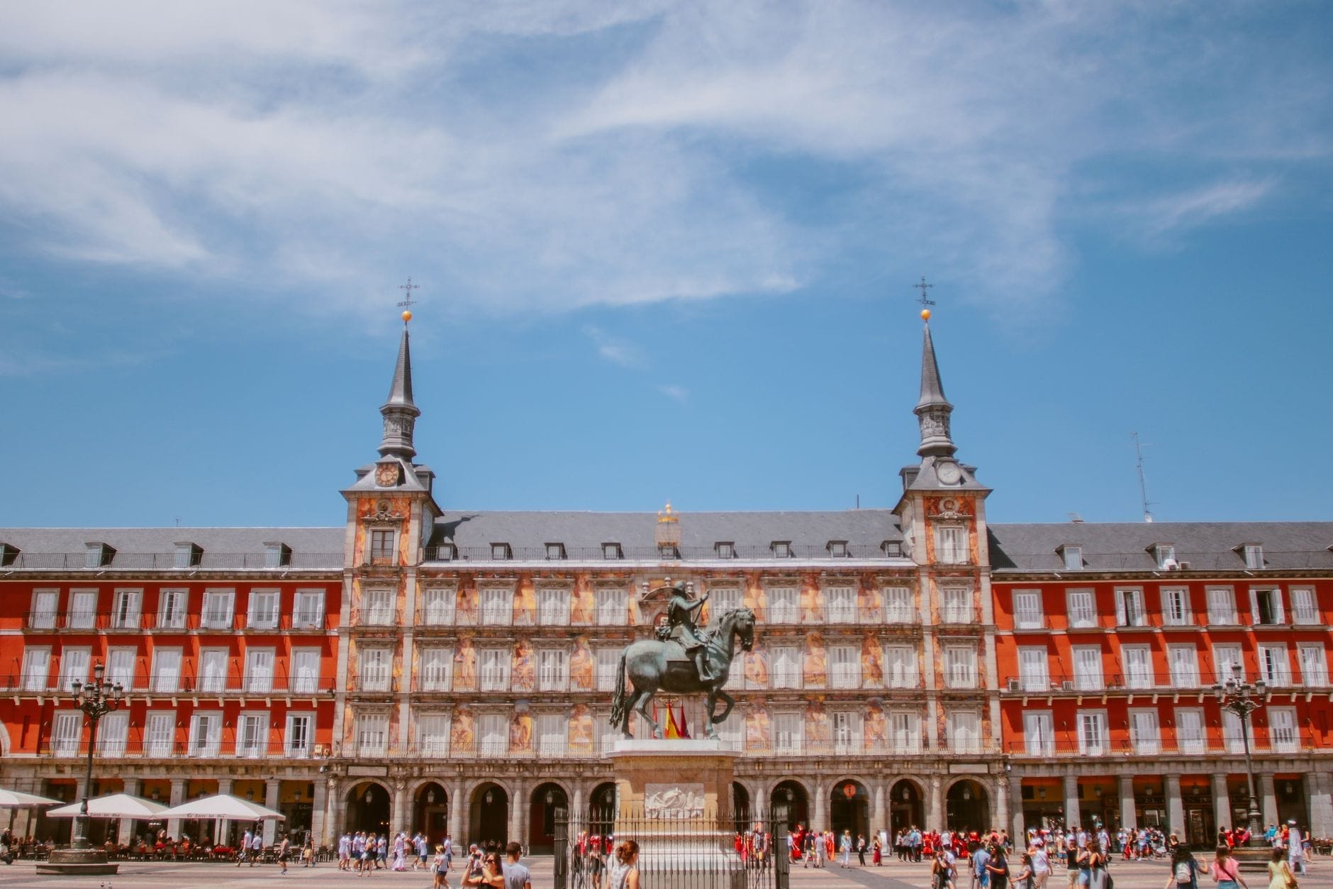 Vista diurna e soleggiata della Plaza Mayor di Madrid con i suoi edifici storici rossi, la statua equestre al centro, il cielo azzurro e la folla animata nella piazza.