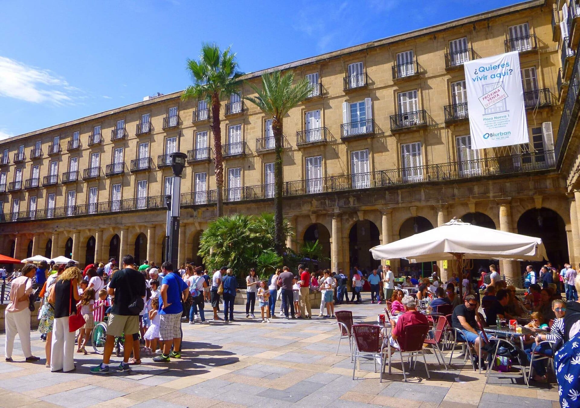 Crowds gather under palm trees and stone arches at Plaza Nueva in Bilbao, with people enjoying outdoor café seating on a sunny day.