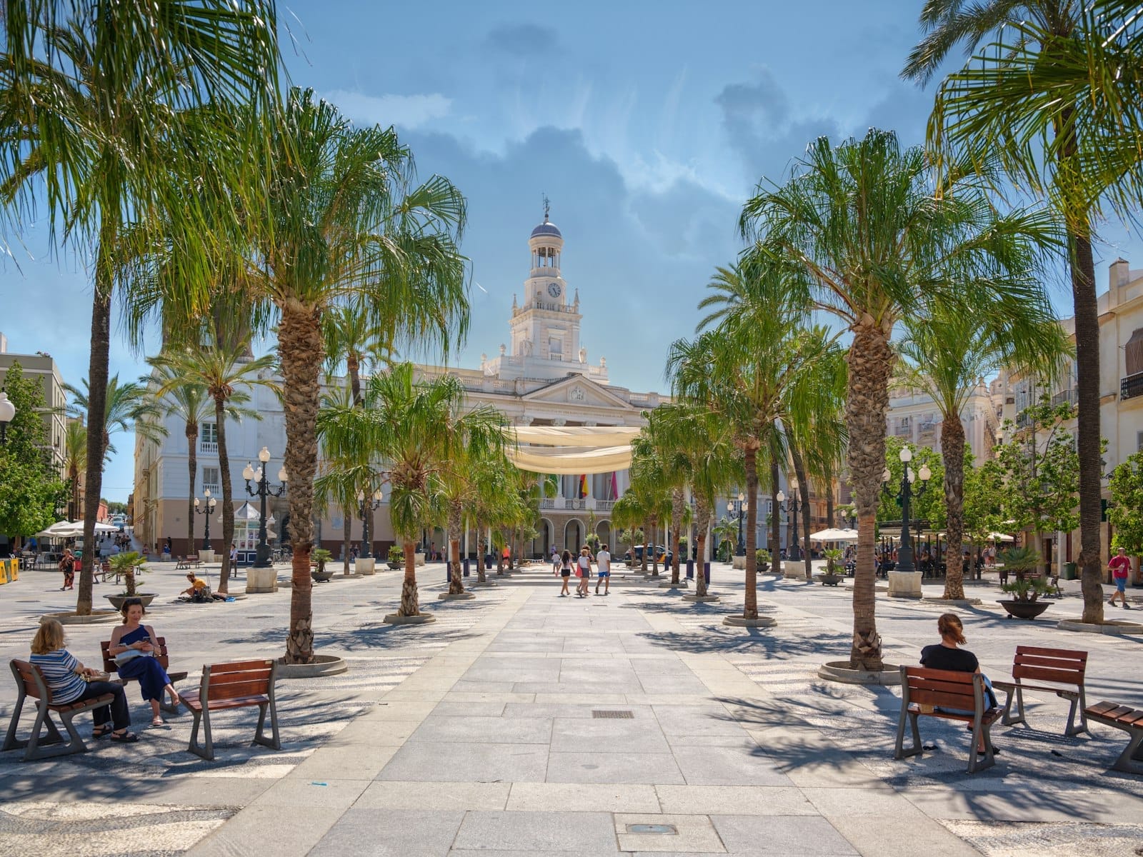 Vista ampia della Plaza de San Juan de Dios a Cádiz con palme, panchine, persone che passeggiano e il palazzo neoclassico del Municipio sullo sfondo.