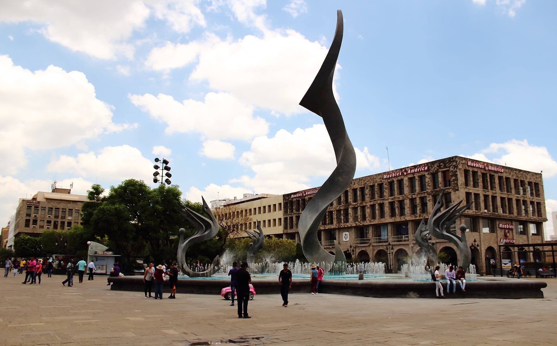 Vista ampia di Plaza Tapatía con sculture metalliche moderne, una grande fontana, edifici civici circostanti e persone che passeggiano sotto un cielo parzialmente nuvoloso.