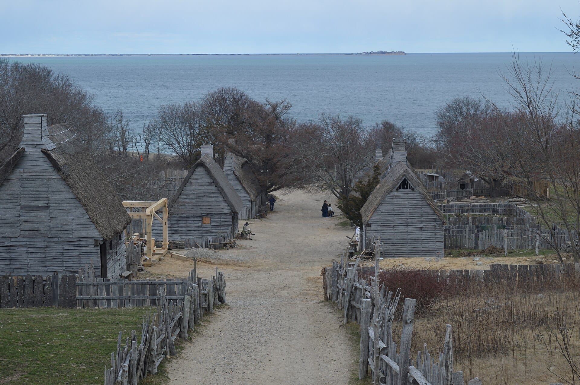 Villaggio storico del Plimoth Patuxent Museums con case in legno consumato, recinzioni in legno spaccato e una vista sull'oceano sotto un cielo nuvoloso.