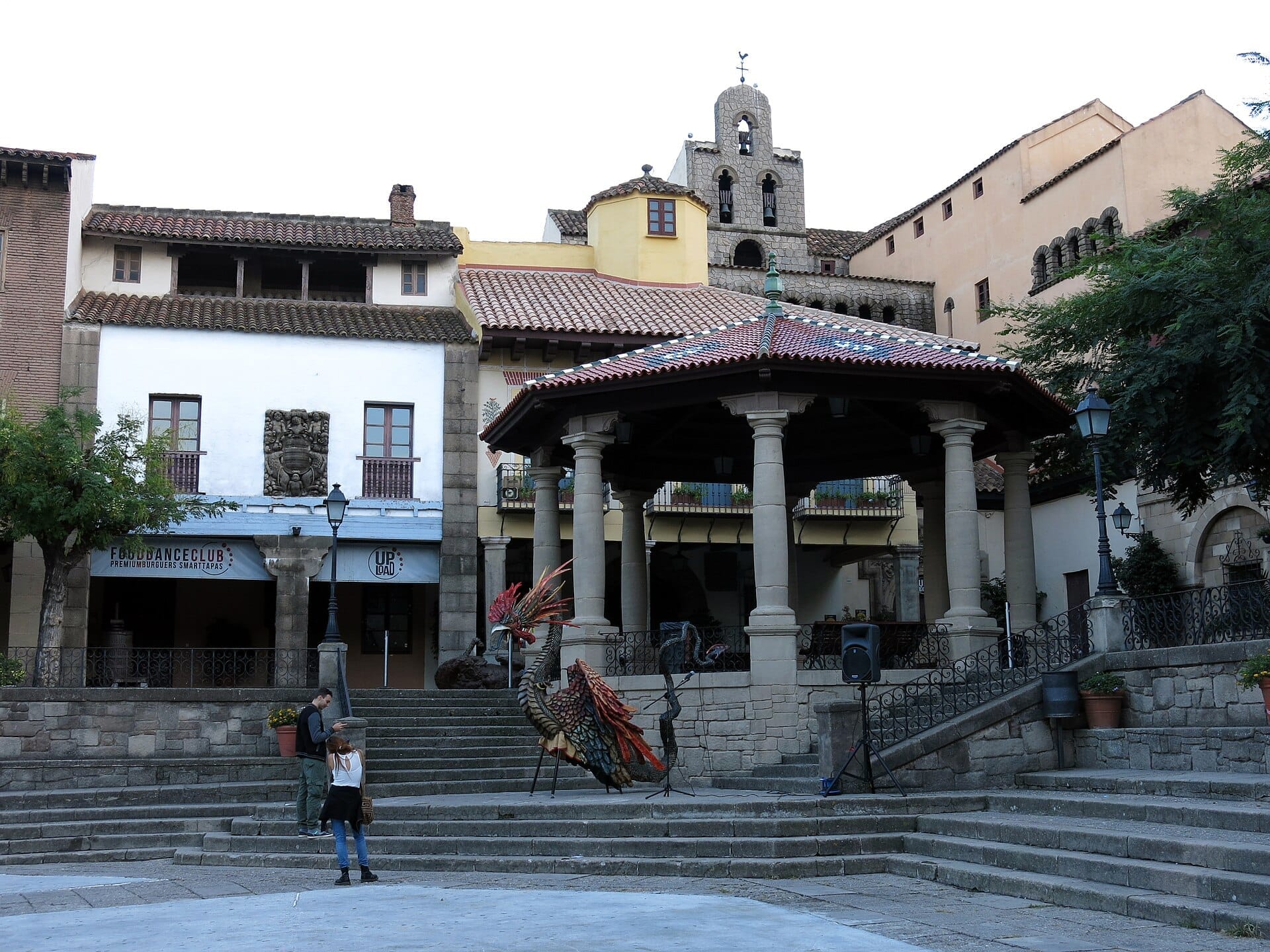 Central plaza at Poble Espanyol, featuring historic Spanish architecture, a colonnaded pavilion, mosaic rooster sculpture, and visitors exploring during daylight.
