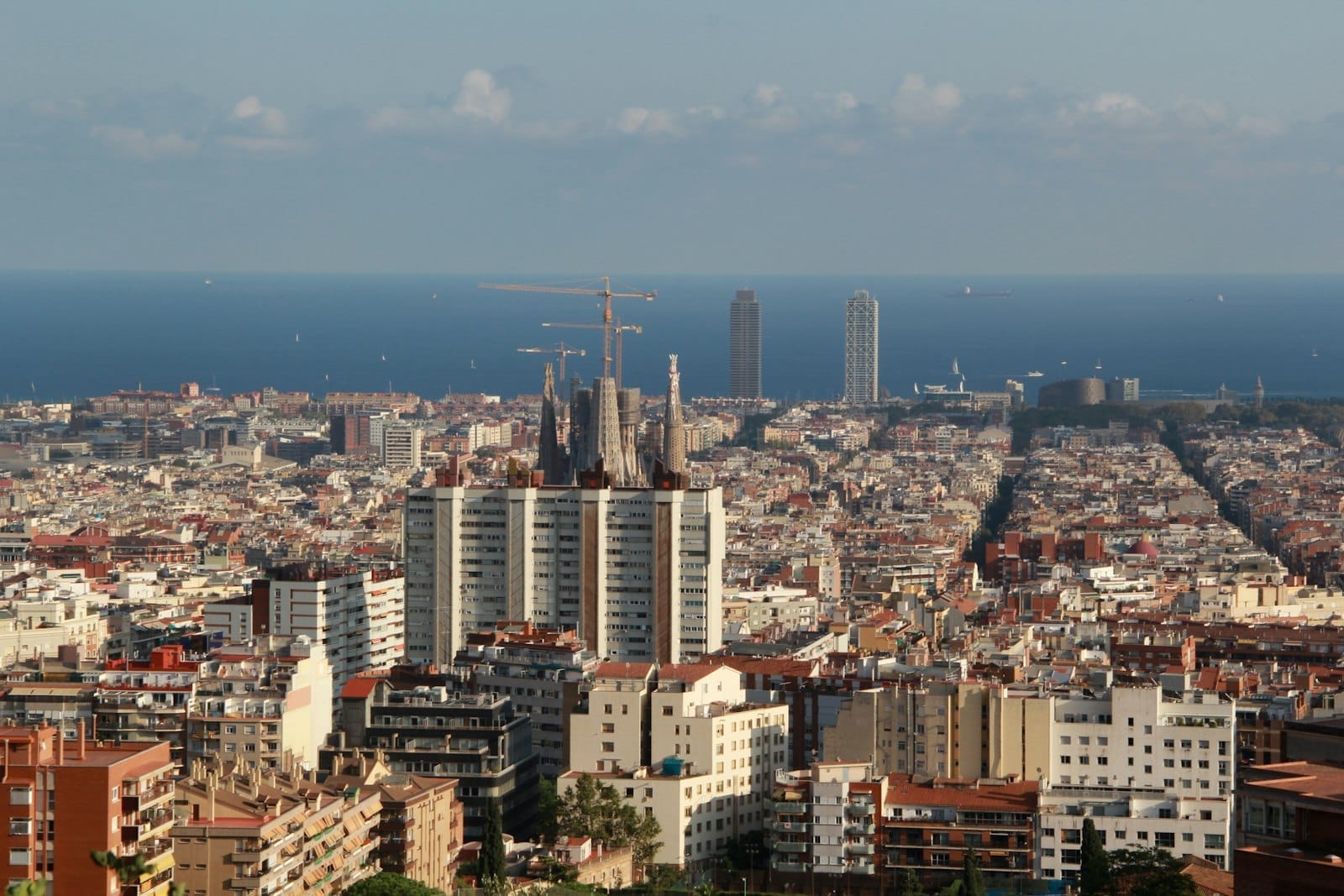 Panoramic view of Barcelona’s Poblenou and Olympic Village with the twin towers, city grid, and Mediterranean Sea under a bright blue sky.
