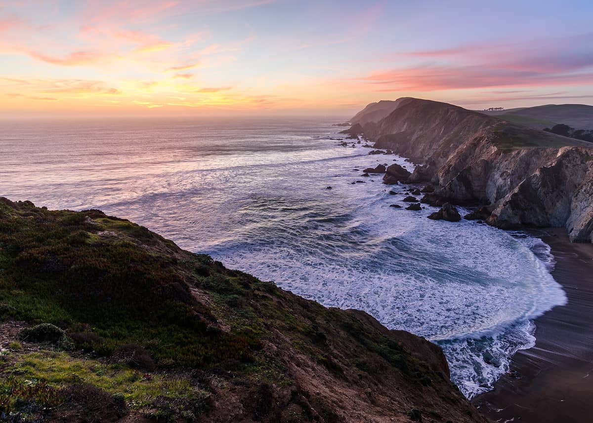 Espectacular atardecer en Point Reyes National Seashore con acantilados escarpados, olas del Pacífico rompiéndose abajo y cielos en tonos pastel sobre la extensa costa.
