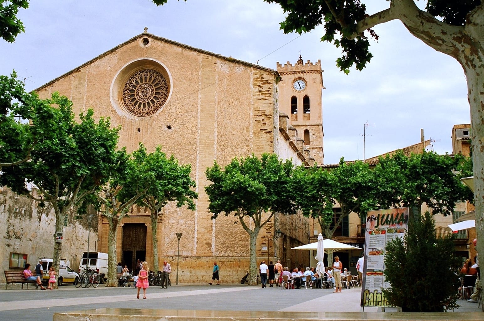 Facciata storica di una chiesa e torre dell'orologio nella città vecchia di Pollença, Mallorca, con piazza alberata, persone che si rilassano e atmosfera soleggiata.