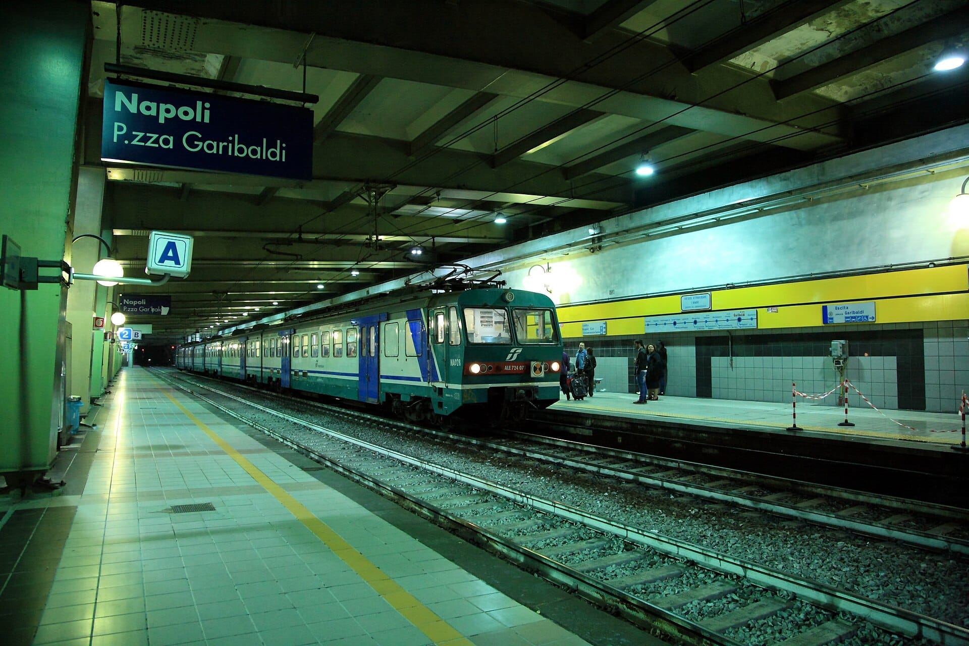 Vista ampla da plataforma da estação de trem Napoli Piazza Garibaldi com um trem da Circumvesuviana, sinalização clara e viajantes esperando ou caminhando.