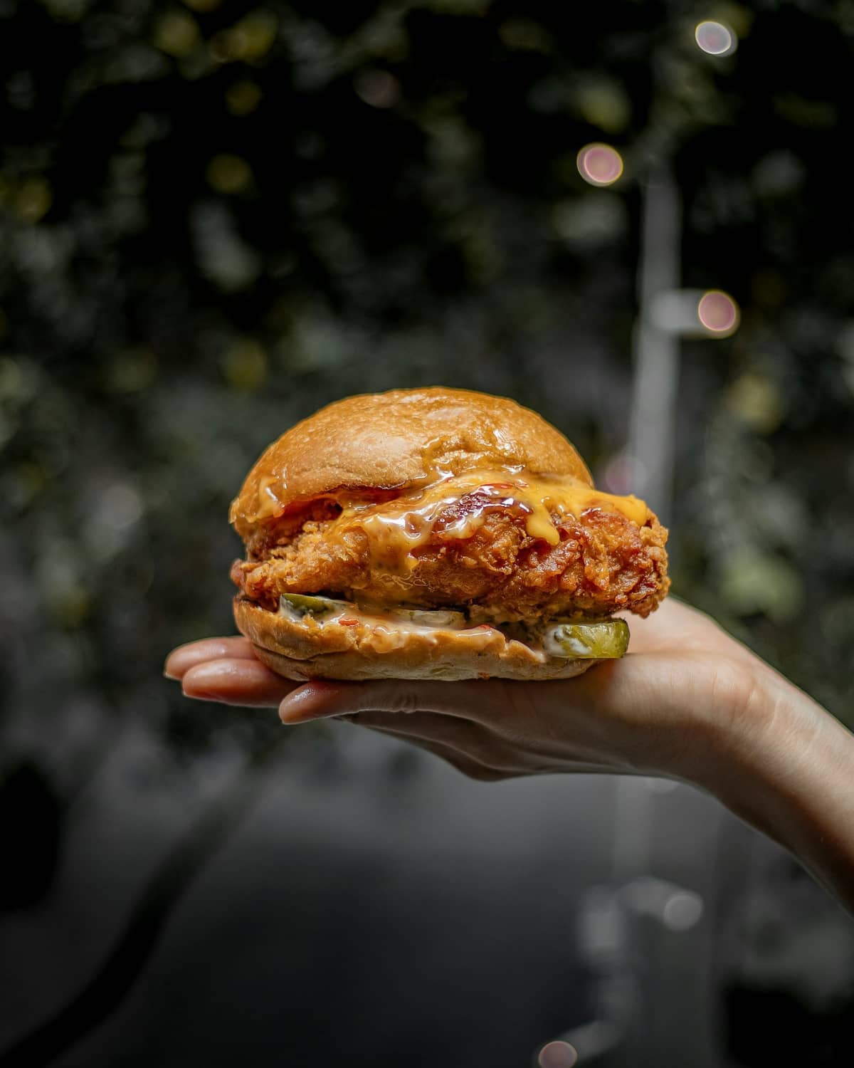 Ein Fried-Chicken-Sandwich, in einer Hand gehalten, in Ponsonby Central in Auckland: Brioche-Brötchen, knuspriges Filet, Gurken und Sauce vor dunklem Hintergrund.