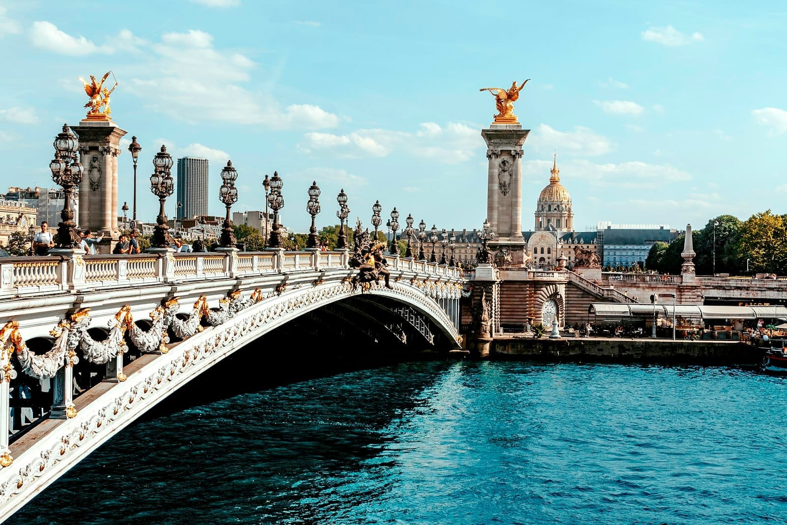 Vista panoramica del Pont Alexandre III in piena luce, con i suoi eleganti lampioni, le statue dorate, l'acqua azzurra e lo skyline di Parigi sullo sfondo.
