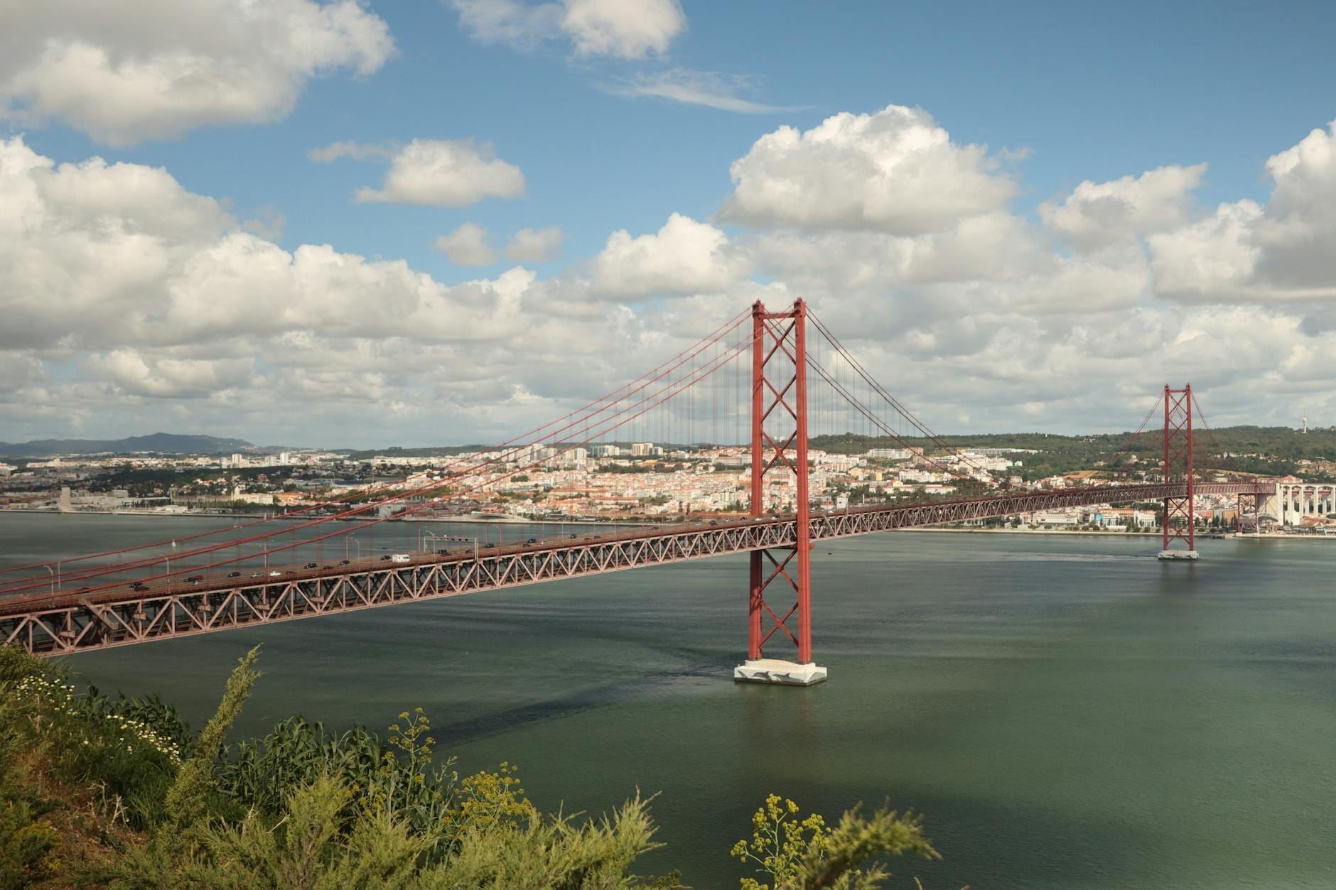 Vista panoramica del ponte sospeso Ponte 25 de Abril che attraversa il fiume Tago, con lo skyline di Lisbona e un cielo azzurro nuvoloso sullo sfondo.
