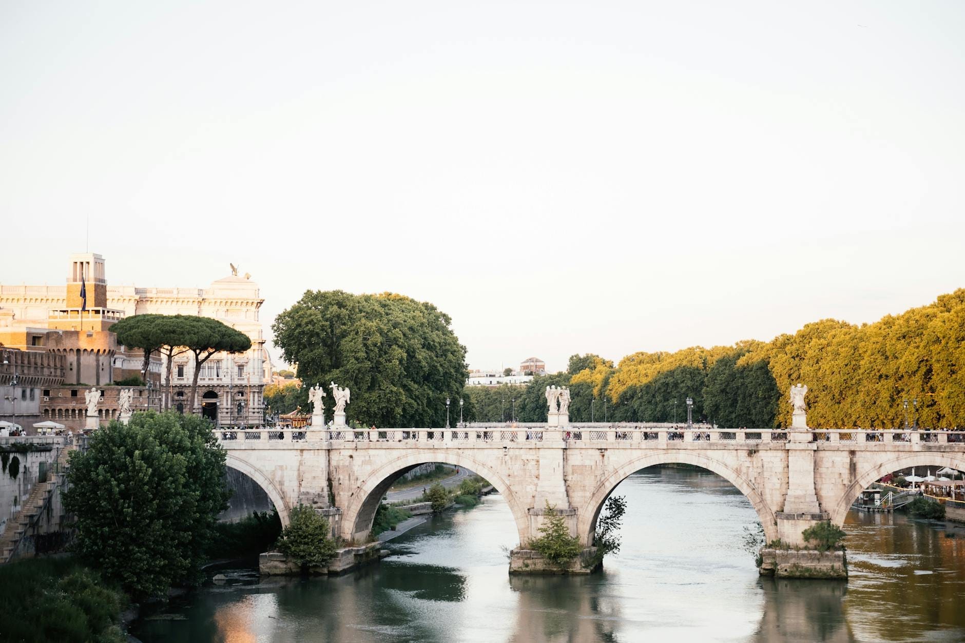 Ponte Sant'Angelo bridge with Baroque angel statues crossing the Tiber River, lush green trees, and historic Roman buildings on a bright, sunny day.