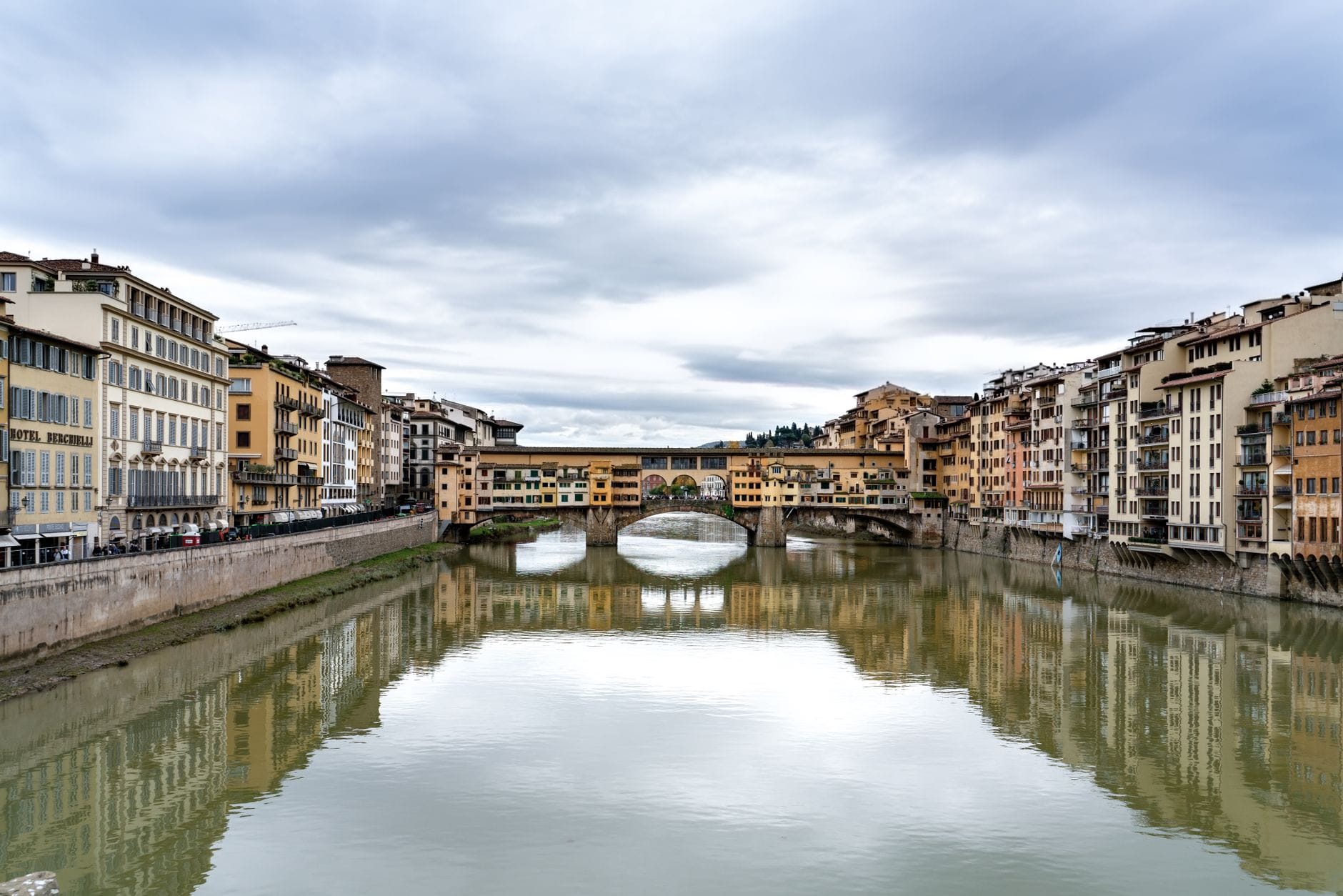 ภาพกว้างมุมสูงของ Ponte Vecchio สะพานประวัติศาสตร์ในฟลอเรนซ์ที่รายล้อมด้วยร้านค้า ข้ามแม่น้ำอาร์โน มีเงาสะท้อนของสะพานและอาคารริมฝั่งน้ำ
