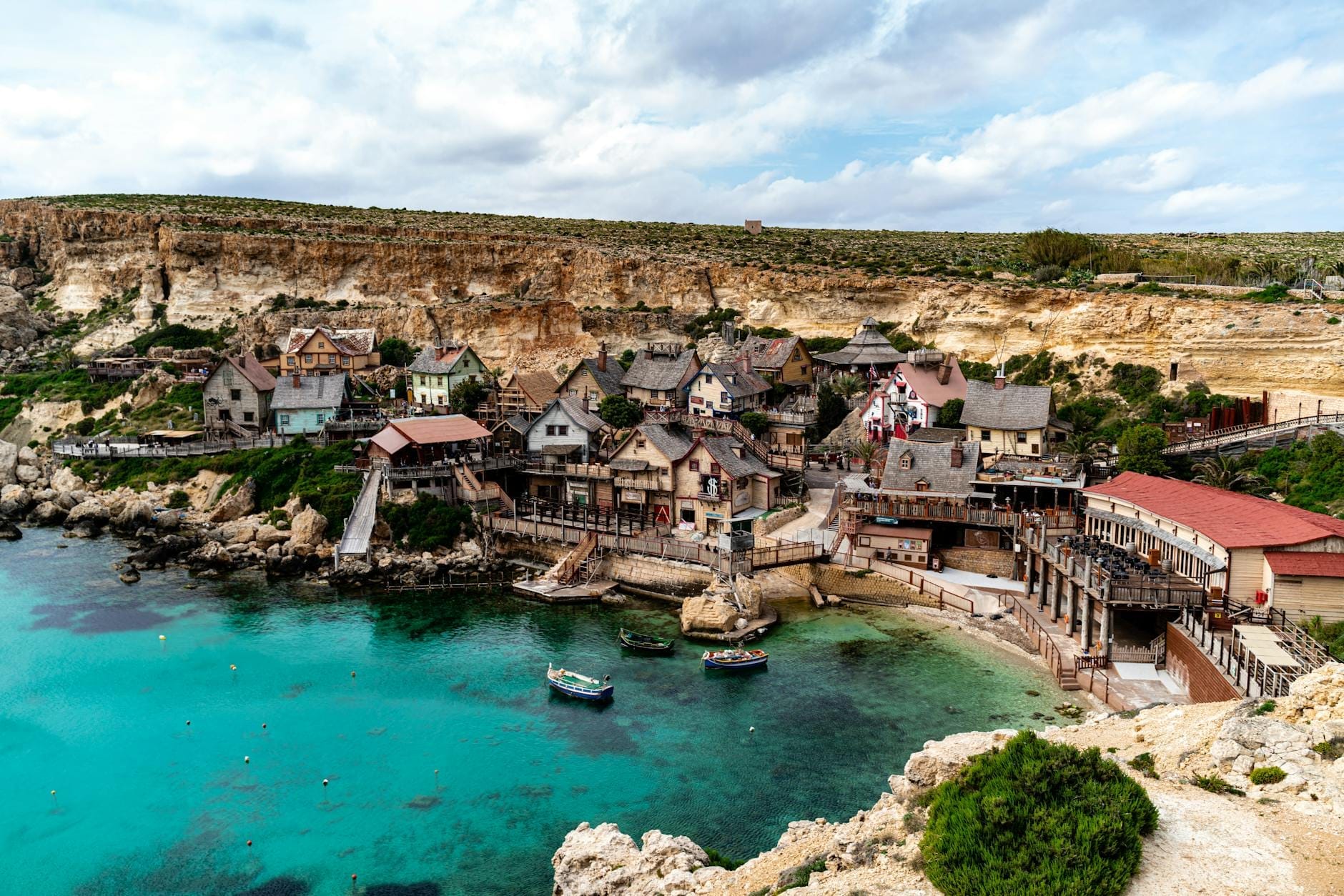 Una vista ampia e colorata delle stravaganti casette in legno di Popeye Village affacciate sulla turchese Anchor Bay, con le scogliere sullo sfondo sotto un cielo luminoso.